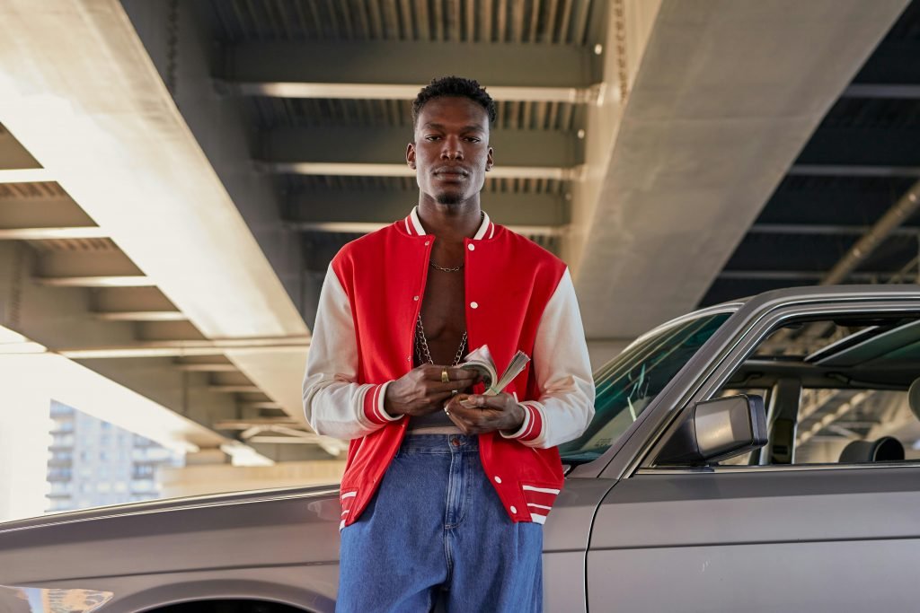 Stylish young man in red jacket holds cash while leaning on a gray car under a bridge, urban setting.